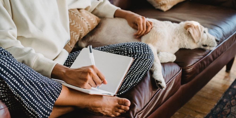 Blogbeitrag zum Thema entspanntes Schreiben - Bild zeigt eine Dame mit Hund, die auf dem Sofa sitzt und entspannt in einem Notizbuch schreibt.